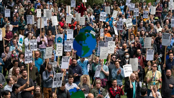 A group of approximately 100 protesters holding climate change protest signs stand outside, with a large cardboard globe in the middle of the group.