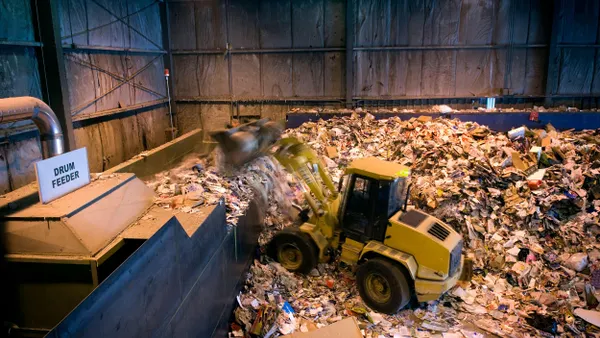 A large yellow front loader moves a scoop of shredded paper and cardboard onto a conveyor labeled "Drum Feeder" inside a dimly lit industrial warehouse. The floor and surrounding area are covered in piles of mixed paper waste and recyclables.