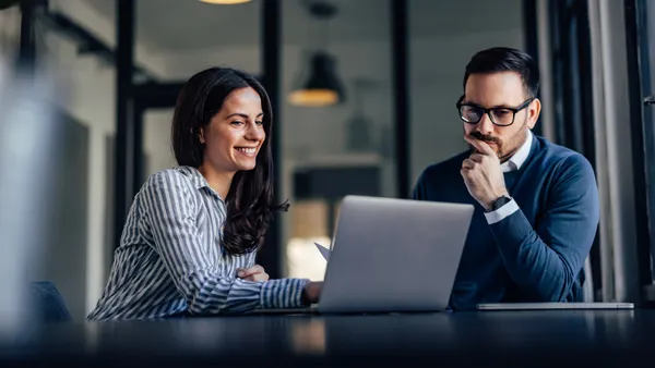 A woman and man sit at a desk, focused on a laptop. She smiles while he gazes thoughtfully. The office setting is modern with soft lighting.