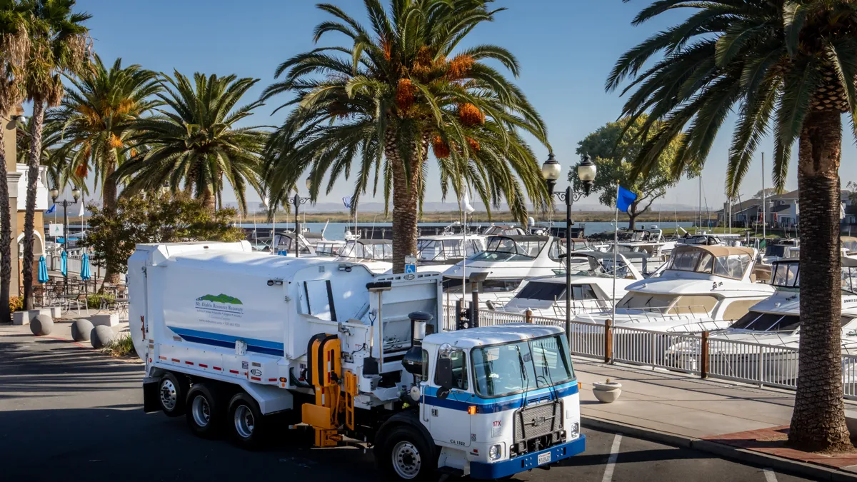 A side-loading collections vehicle is parked on a road in front of a line of palm trees and marina filled with boats.