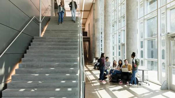 Two students walk down a staircase inside a campus building while another group students gather in the corridor.