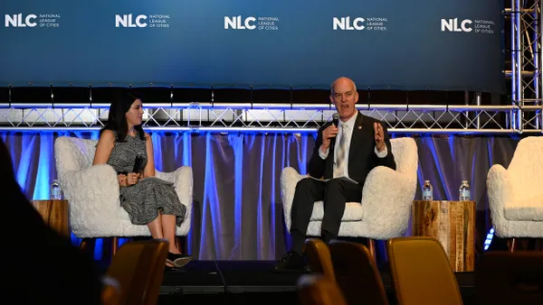 Woman and man seated on a stage in front of a banner that reads "NLC - National Leage of Cities."