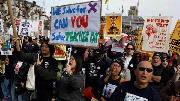 A large group of protesters stand together holding signs. One large sign in the center of the crowd says "We solve for X. Can you solve teacher pay?"
