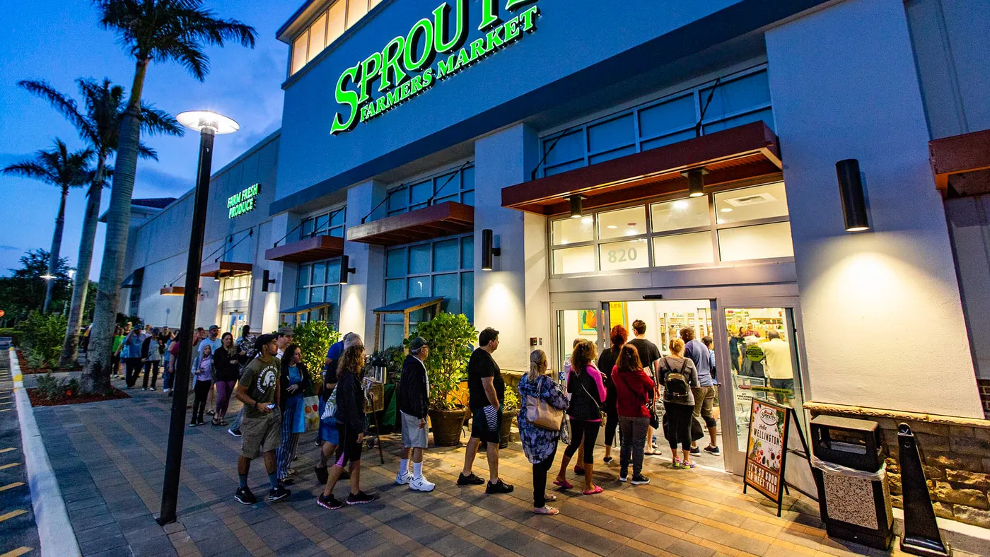 A line of people wait to enter a grocery store, a sign reads "Sprouts."