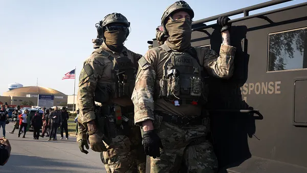 Federal law enforcement agents in uniform with masks covering their faces ride an armored vehicle through demonstrators during a protest outside an immigrant processing and detention center.