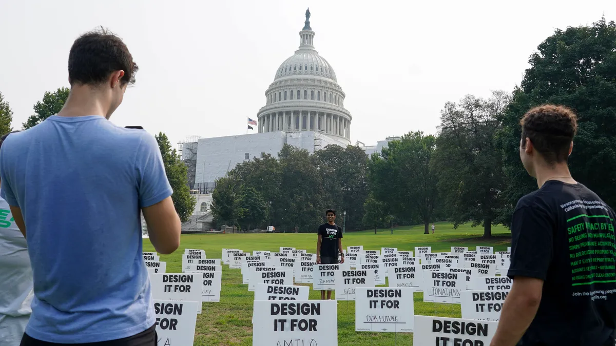 Lawn signs are set up on the lawn near the U.S. Capitol Building in Washington, D.C. The signs say "Design It For" and the last word is filled in with different names. Three people are looking at the signs.