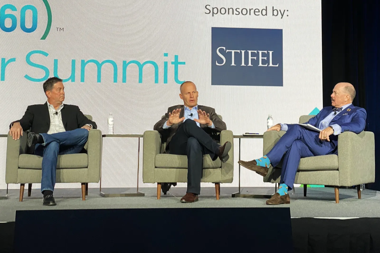 Three people in suits sit on a stage during an event. Behind them, a digital screen notes the event is &quot;Sponsored By: Stifel&quot;