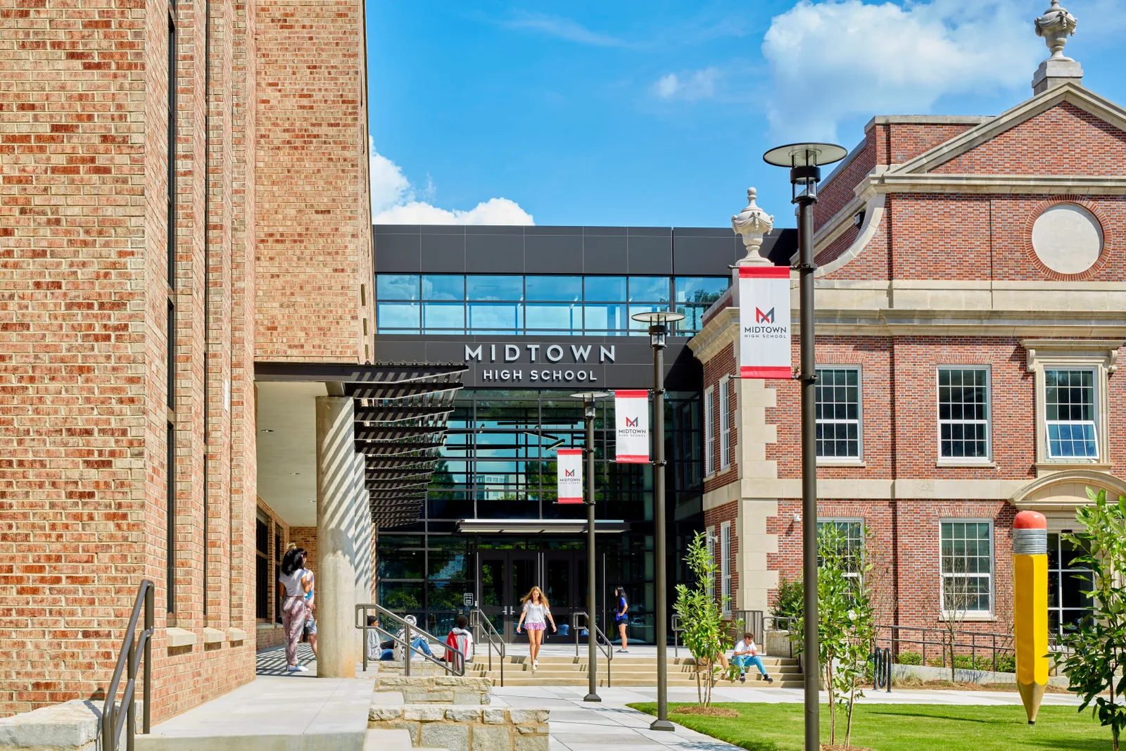 The outside of a school building shows students sitting and walking and a pencil sculpture.