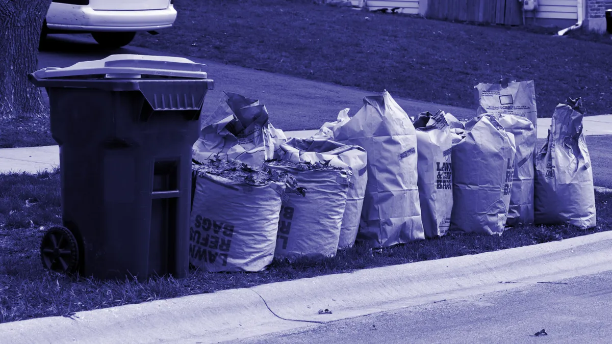 Paper bags filled with leaves and a trash can sit on a grass curb in a residential area.