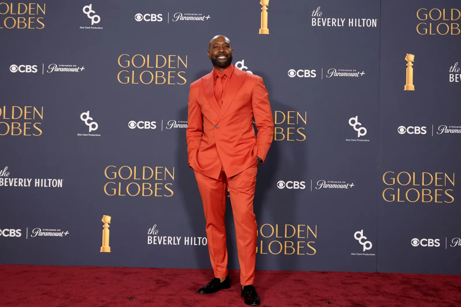 A person in an all-red suit shirt and tie stands in front a sign that reads Golden Globes.