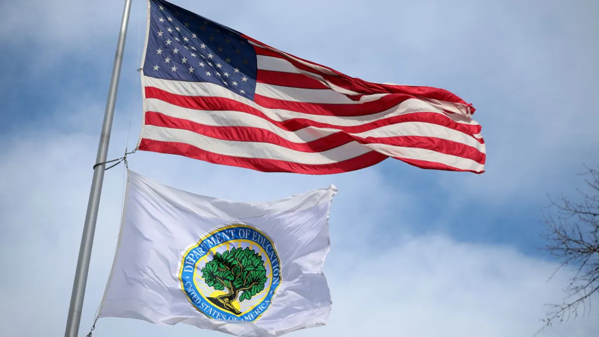 Two flags fly in the wind on a pole outside. The background is a clear blue sky with clouds