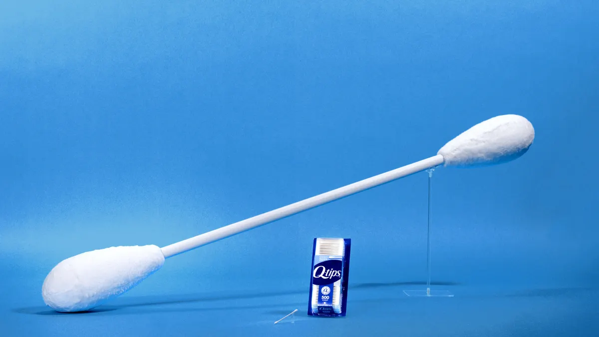 A giant Q-tip stands above a standard package of the cotton swabs against a blue background
