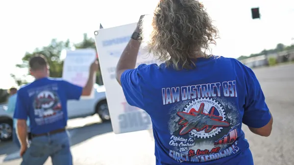The back of a person in a blue t-shirt with the IAM District 837 text and logo and is holding a sign.