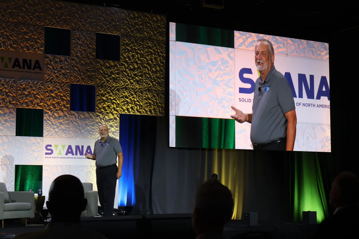 A man wearing a polo stands on a stage with a live feed projected behind him. The stage backdrop is decorated with the SWANA logo.