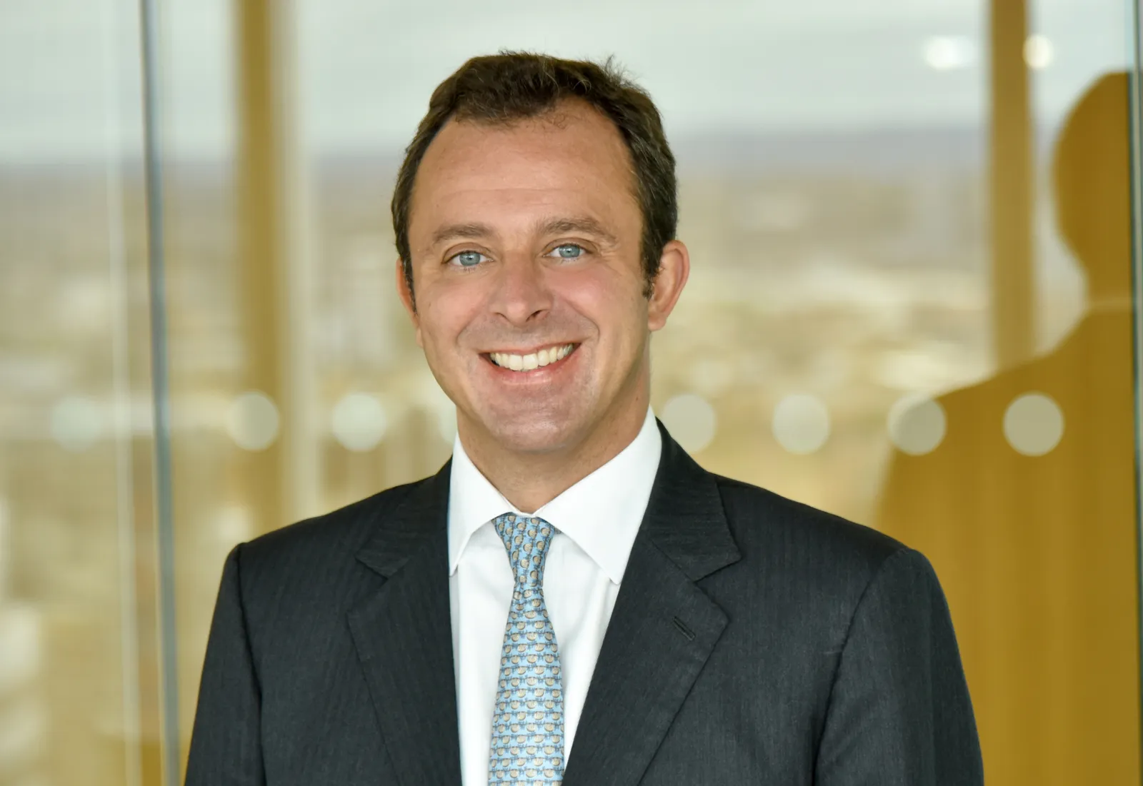 Jan Bellens, wearing a dark suit and blue tie, stands in front of a glass panel