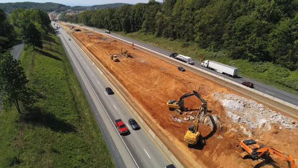 Construction heavy machinery moves along dirt next to two roads that stretch out of frame on a clear, sunny day.