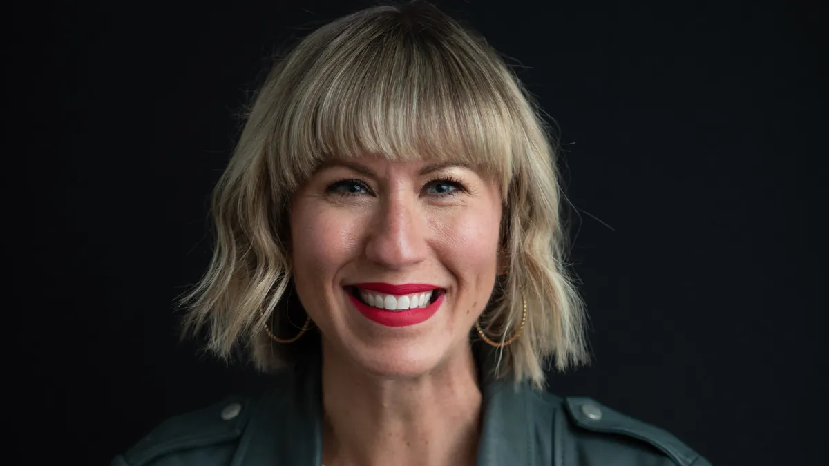 A headshot of a person with bobbed blond hair and bangs, wearing red lipstick and smiling.