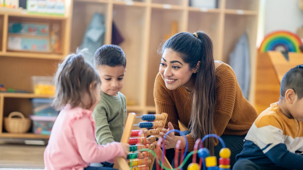 A female Kindergarten teacher sits on the floor with students as they play with various toys and engage in different activities.