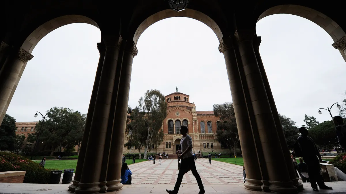 A student walks in front of arches, with a campus building in the background, on a college campus.