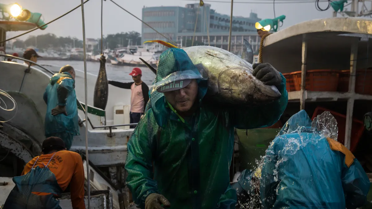 A fisherman with his hood up unloads fish as other fishermen stand in the background near a boat