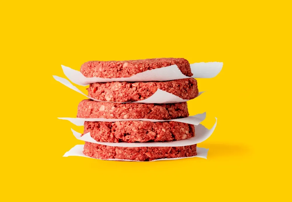 A stack of uncooked Impossible Burger patties separated by wax paper on a yellow background