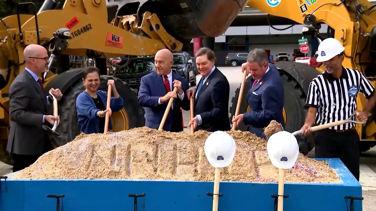 Six people stand behind a box of dirt with shovels and construction equipment in the background.