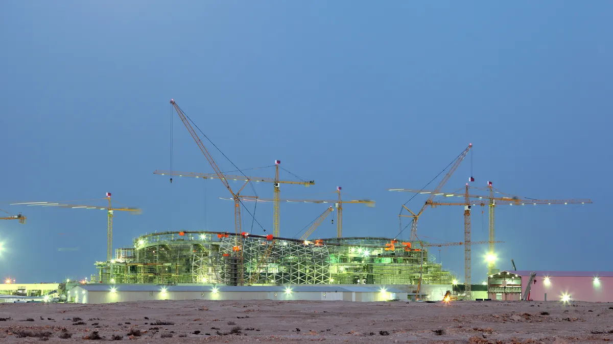 Several cranes tower over the frame of a stadium construction site in the desert.