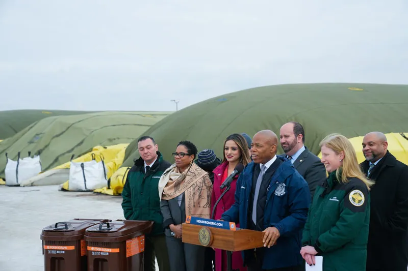 Several dignitaries stand around a lectern in front of large mounds covered in tarps.