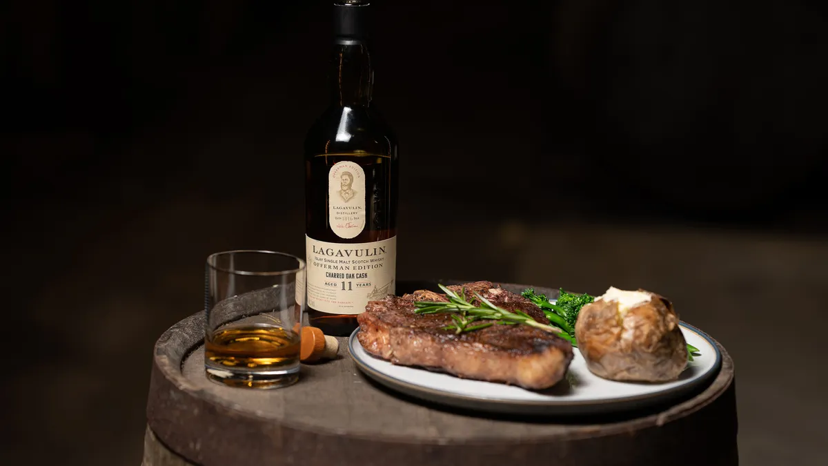 A bottle of whiskey sits on a cask next to a plate of steak.