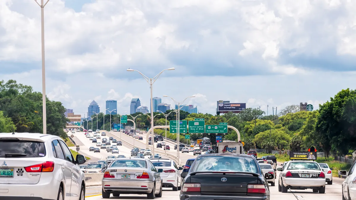 Passenger cars are several lanes of highway with a skyline of city buildings during a sunny day in the background.