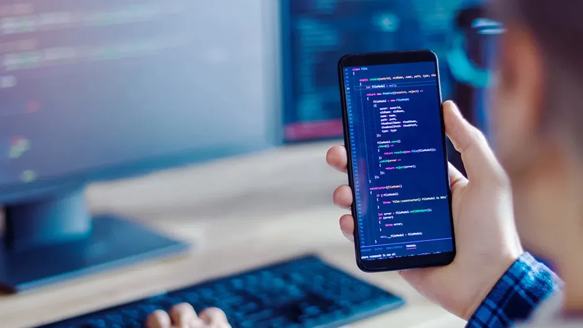 A photo of a hand holding a cell phone with code on it, with a computer on a desk in the background of the picture.