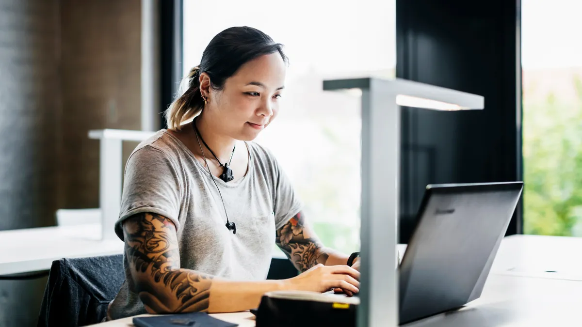 An office worker with tattoos works on a laptop at their desk.