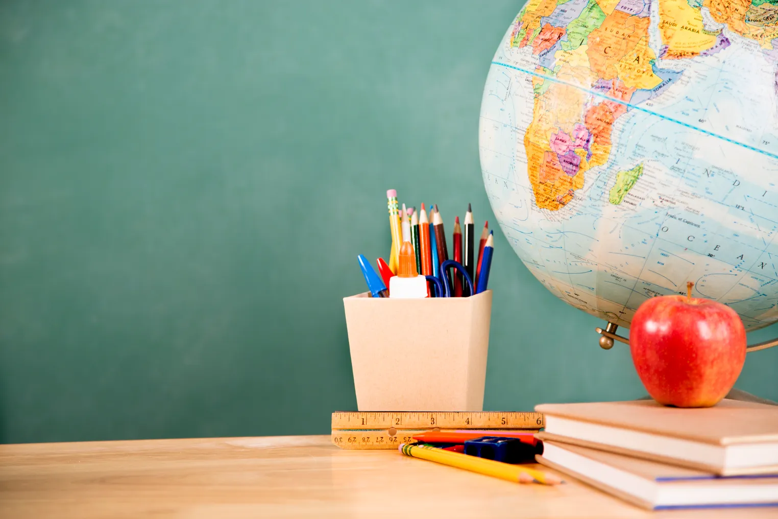 A world globe sits on a desk against a chalkboard background. A red apple, books, ruler and pens and pencils in a pencil holder also sits on the desk.