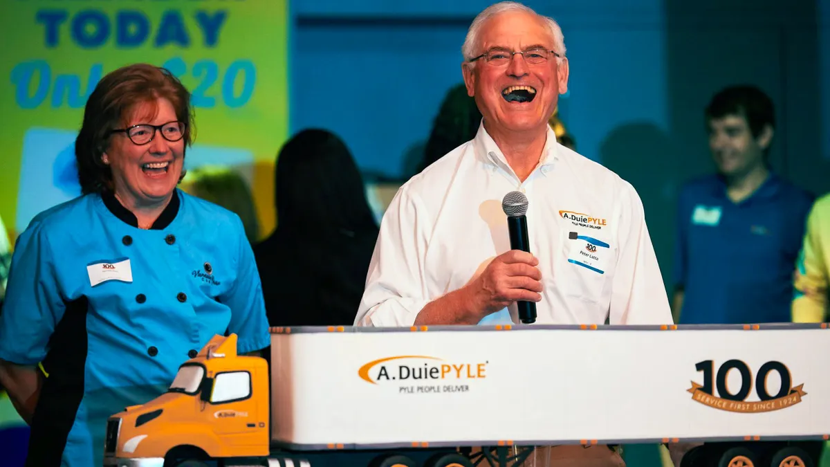 Two people laugh behind a replica cake of an A. Duie Pyle truck, the trucks trailer reads, “100 service since 1924”.