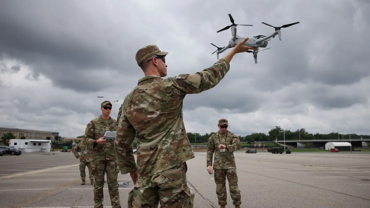 Three U.S. Army soldiers in green and brown camouflage uniform, with one holding a drone in one hand.