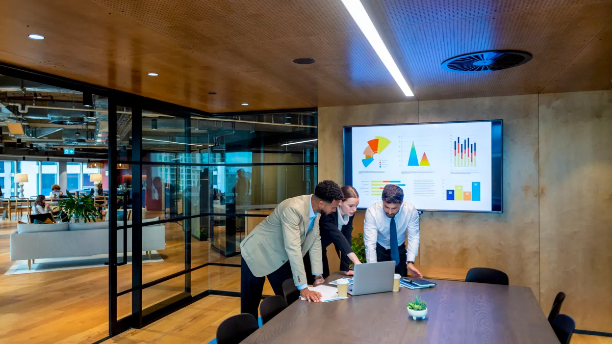 Business people working on a laptop computer in a modern office board room. There is paperwork on the table with charts and graphs on it. There is a screen behind them with financial charts and graphs