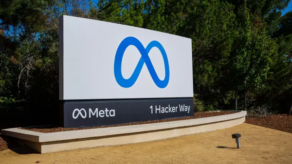 A large outdoor sign features Meta's blue infinity loop logo on a white background above the company name "Meta" and the address, "1 Hacker Way." The sign sits on a curved concrete base in a landscaped area with mulch and dirt.
