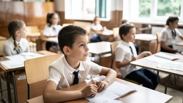 A student is sitting at a desk in a classroom. There are papers on the desk and the student is looking toward the front of the classroom. There are other students in the room.