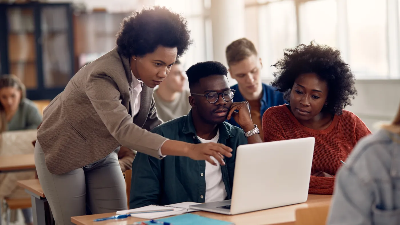 A Black woman helps two Black young adults who are seated in front of a laptop computer.