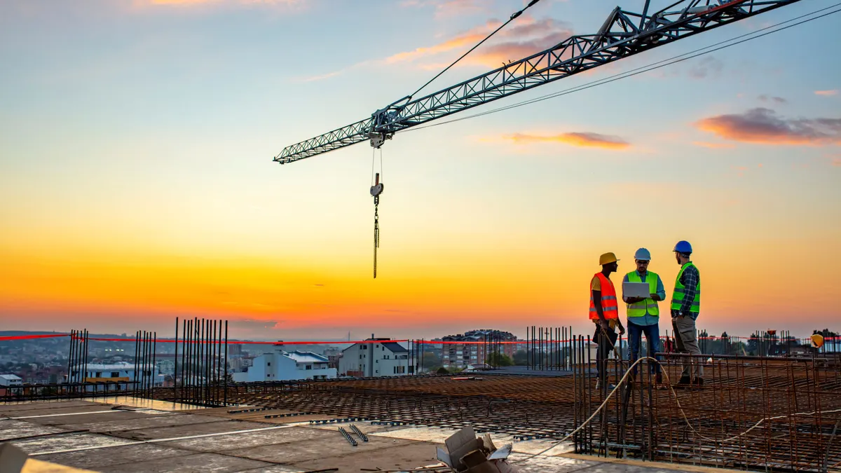 Three construction workers talk under a crane on a jobsite's roof.