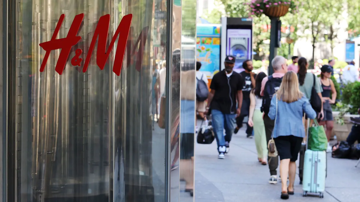 A red H&M logo is seen on a mirrored store exterior as people walk by.
