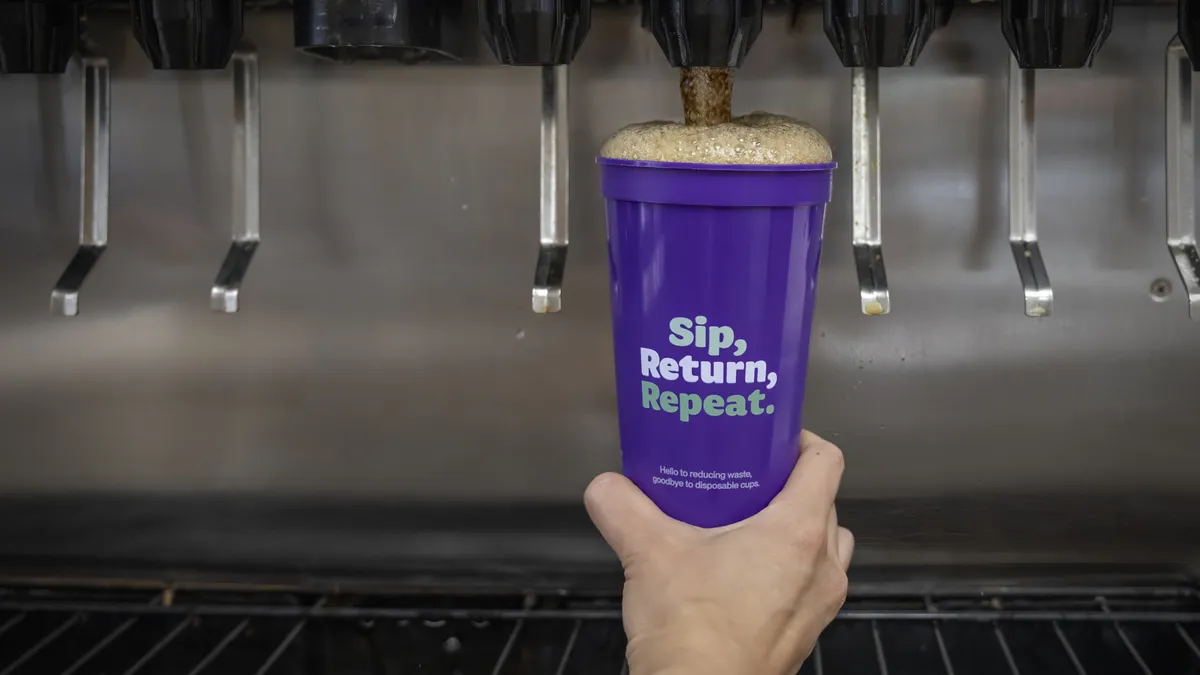 A hand holds up a reusable cup being filled by a soda fountain. The purple cup says "Sip, Return, Repeat."