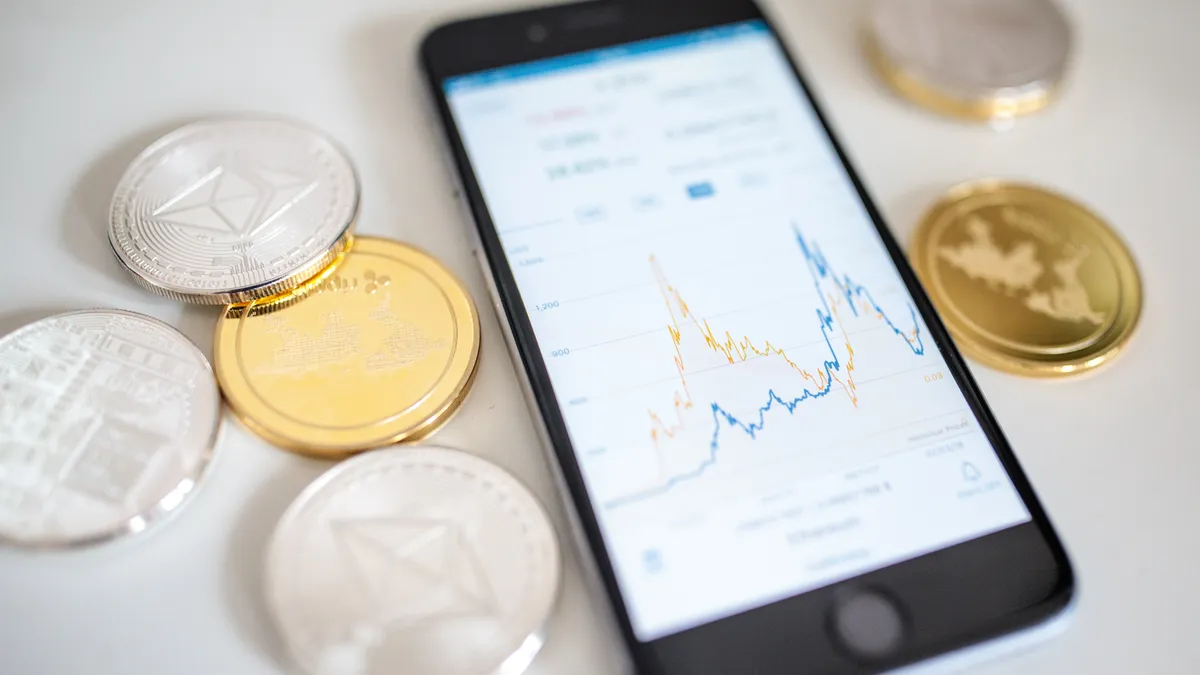 Cryptocurrencies laying on a flat surface next to a smartphone that shows a chart with zig-zagging lines.