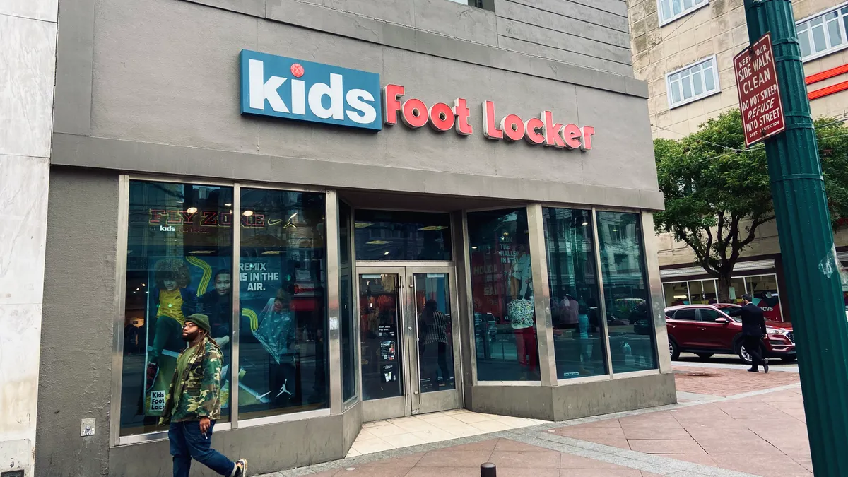 A person wearing a camo jacket walks by a gray granite storefront that says "kids" in a blue square and "foot locker" in red letters.