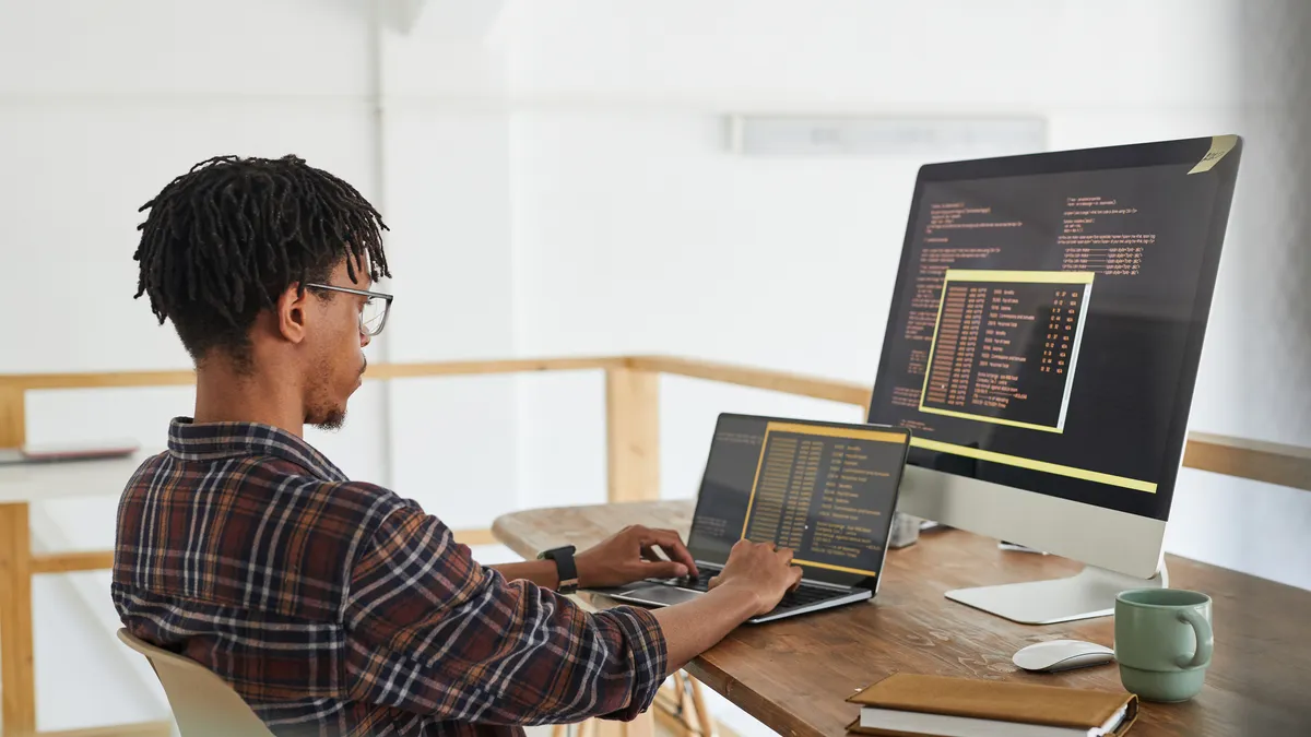 African-American IT developer typing on keyboard with black and orange programming code on computer screen and laptop in contemporary office interior, copy space