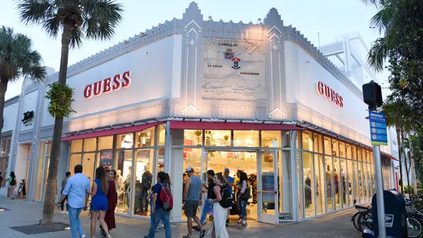 Outside corner view of a Guess store, with a white facade, palm trees lining the street, and crowds of shoppers milling about.