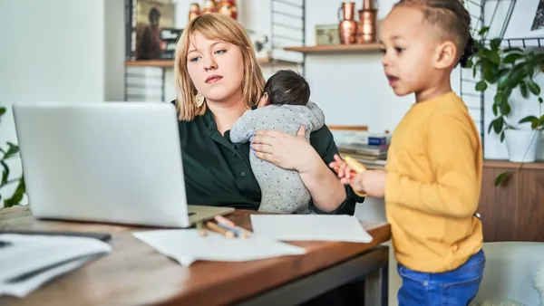 Single mother carrying newborn baby and working at home. Woman using laptop while son playing in living room.