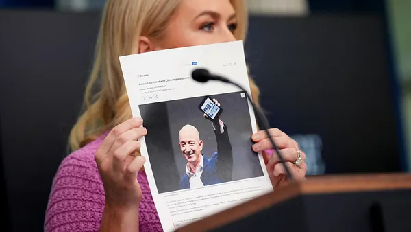 A person with long blond hair, wearing a pink shirt, holds up a photo of another person.