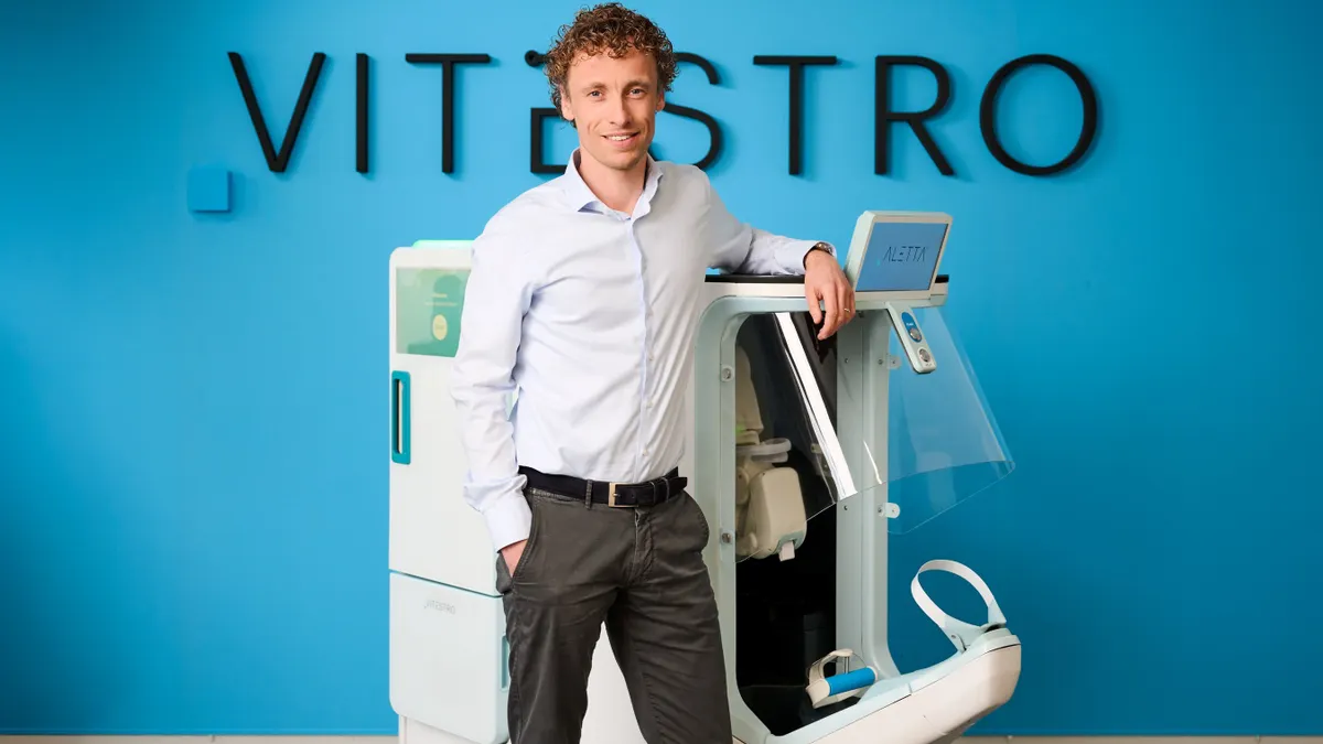 A man wearing a white shirt and grey pants stands in front of a robotice blood drawing machine.
