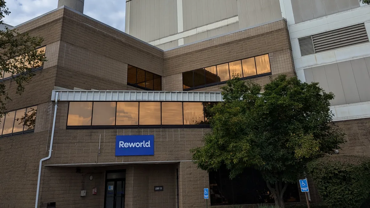 Entrance to a building with reflective windows leading to a larger industrial shed. A smokestack is visible behind the building, as well as a sign on its front reading “Reworld.”